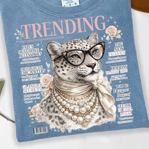 Könnte beinhalten: Ein blaues T-Shirt mit einem Aufdruck eines Leoparden, der eine schwarze Brille, eine Perlenkette und ein Seidentuch trägt. Das Motiv ähnelt einem Magazin-Cover mit dem Text TRENDING, verschiedenen Schlagzeilen und kleinen Blumenmotiven.