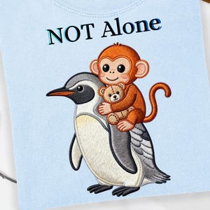 May include: Light blue t-shirt with the text "NOT Alone" above an embroidered design. The design features a cartoon monkey holding a teddy bear, riding on the back of a penguin.