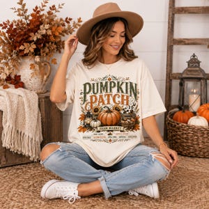 May include: An off-white short-sleeve t-shirt featuring a graphic print with the text PUMPKIN PATCH FARM MARKET. The design includes illustrations of pumpkins, sunflowers, a lantern, and autumn leaves, along with smaller text listing seasonal items.