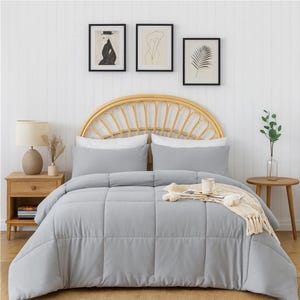 May include: A light gray comforter set on a bed with white pillows and a woven headboard. The bed is in a room with framed art and a wooden side table with a lamp. A cream-colored throw blanket and a mug are on the bed.