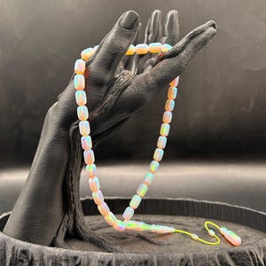 Puede incluir: Un rosario de cuentas cilíndricas iridiscentes con aspecto de ópalo que emiten un suave resplandor. Las cuentas están unidas por un cordón fino de color amarillo brillante, que termina en una pieza cilíndrica a juego con una pequeña lazada.
