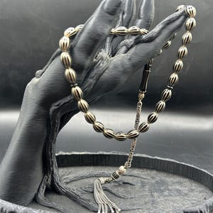 Può includere: Una corona di preghiera con perline ovali a righe bianche e nere. La collana include un separatore in metallo argentato, un distanziatore decorativo argentato e una nappa argentata all'estremità, creando un design dettagliato e strutturato.