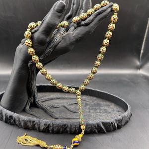 Peut inclure: Un chapelet composé de perles sphériques dorées ornées de motifs géométriques complexes en émail rouge, bleu et vert. Le cordon comprend une perle intercalaire cylindrique, une perle en verre décorative et un pompon de couleur dorée.