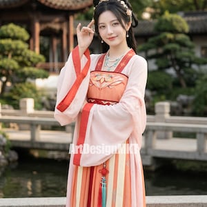 Puede incluir: Una mujer con un vestido hanfu chino tradicional. El vestido es principalmente rosa claro con detalles rojos y naranjas. Las mangas son largas y fluidas, y la falda presenta rayas verticales de varios colores. El atuendo incluye un tocado y joyas decorativas.