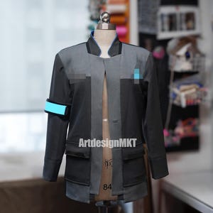 Puede incluir: Una chaqueta de color gris oscuro y gris claro que presenta un cuello alto con ribete azul, un brazalete azul brillante en la manga derecha y un pequeño emblema rectangular azul en el pecho izquierdo. La chaqueta se exhibe en un maniquí.