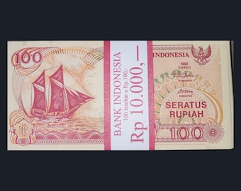 Op de afbeelding: Een stapel Indonesische 100 Rupiah-bankbiljetten, gebonden met een rood-witte papieren band. De bankbiljetten tonen een zeilschipillustratie en de tekst "BANK INDONESIA" en "SERATUS RUPIAH".