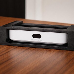 Peut inclure: Un boîtier de montage en rack noir contenant un appareil blanc avec une découpe ovale noire. L'appareil porte l'inscription "Cloud Gateway Ultra". Le support de rack est posé sur une surface en bois.