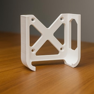 Pode incluir: Um suporte branco impresso em 3D com um design geométrico aberto. O suporte apresenta uma estrutura de suporte em forma de X e vários orifícios circulares para montagem. Está sobre uma superfície de madeira.