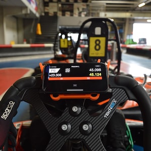 May include: A black smartphone displaying racing data is mounted on the steering wheel of a go-kart. The steering wheel is black with carbon fiber accents and the phone is held in an orange bracket. The background shows a race track.