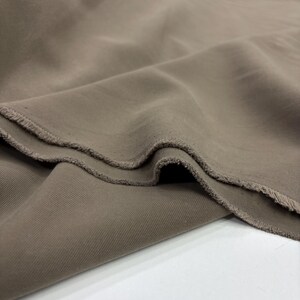May include: A length of solid taupe-colored fabric with a visible diagonal twill weave pattern. The material is draped in soft folds, showcasing a matte finish and raw, slightly frayed edges along the cut sections of the textile material.