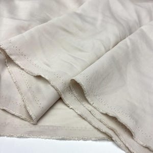 May include: A length of off-white, solid-colored fabric with a visible twill weave texture. The material features raw, frayed edges along the sides, which display a distinct row of small, evenly spaced pinholes running parallel to the selvage.