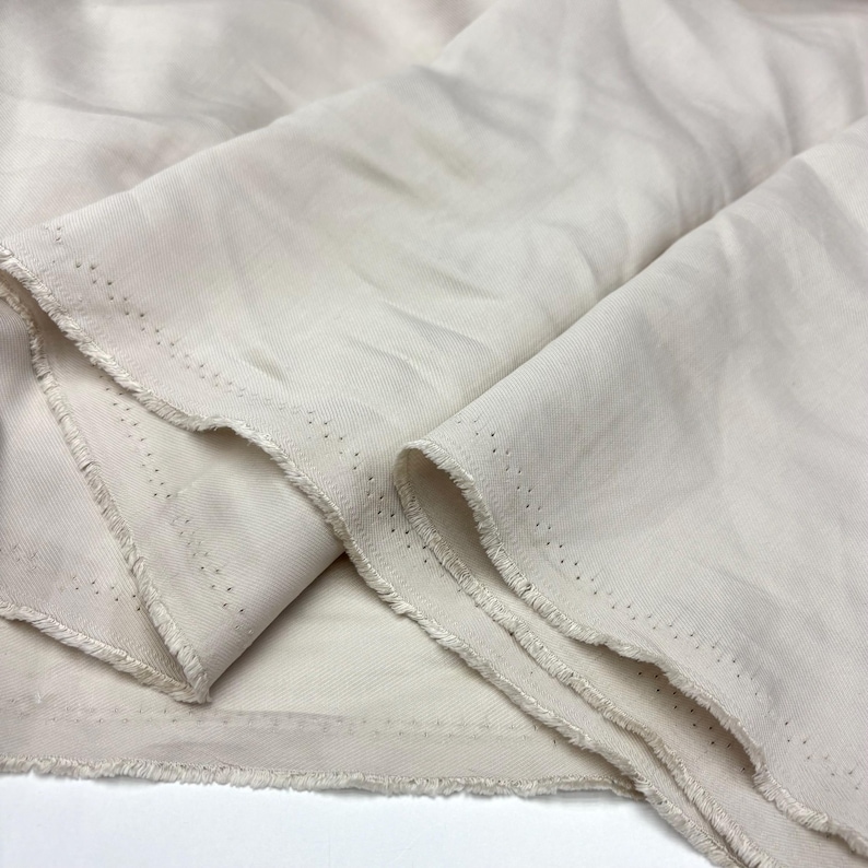 May include: A length of off-white, solid-colored fabric with a visible twill weave texture. The material features raw, frayed edges along the sides, which display a distinct row of small, evenly spaced pinholes running parallel to the selvage.