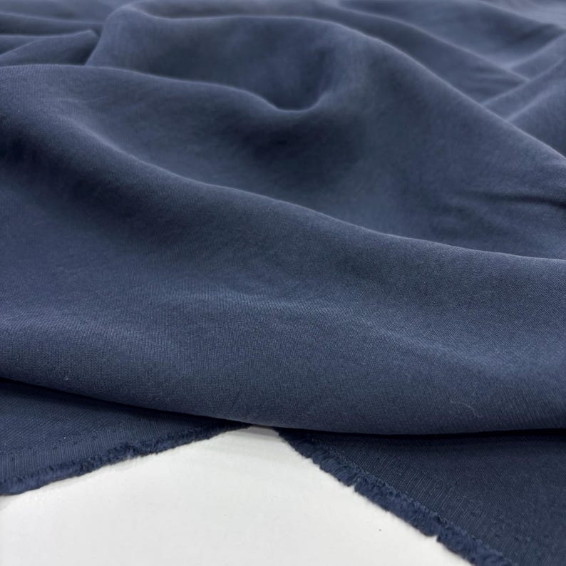 May include: A piece of solid navy blue fabric with a soft, matte texture and visible folds. The material features a slightly uneven, natural weave pattern throughout, with raw, unfinished edges visible at the bottom corners of the textile.