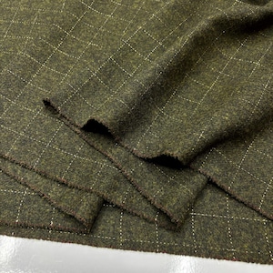 Può includere: Primo piano di tessuto verde oliva con un motivo a rombi creato da cuciture bianche. Il tessuto ha un aspetto strutturato e un bordo grezzo, suggerendo che possa essere utilizzato per abbigliamento o tappezzeria.