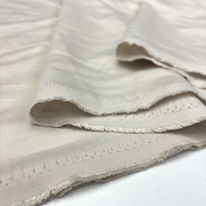 May include: A length of off-white fabric featuring a subtle, diagonal twill weave pattern. The material is folded to show its texture, with visible raw, frayed edges along the selvage that display a series of small, evenly spaced pinholes.