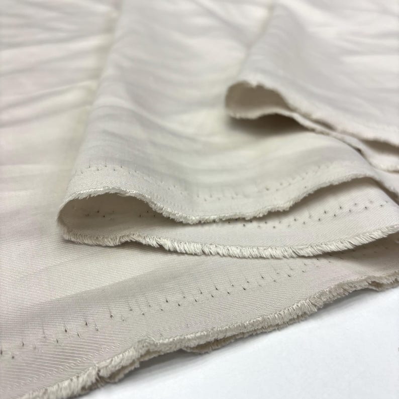May include: A length of off-white fabric featuring a subtle, diagonal twill weave pattern. The material is folded to show its texture, with visible raw, frayed edges along the selvage that display a series of small, evenly spaced pinholes.