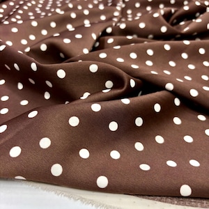 May include: Close-up of a brown fabric with a repeating pattern of white polka dots. The fabric is draped, showing folds and shadows. The material appears to be smooth and possibly satin or silk.