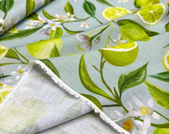 May include: Fabric with a light blue background featuring a pattern of lemons, lime slices, green leaves, and white blossoms. The fabric is folded, showing the raw edge of the material. The design has a watercolor style.