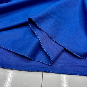 Puede incluir: Primer plano de una tela azul vibrante, posiblemente un textil o material de ropa. La tela est&aacute; doblada, revelando capas y un reverso azul m&aacute;s claro. Se ven costuras blancas en los bordes, lo que sugiere una costura terminada. El material parece liso y posiblemente tejido.