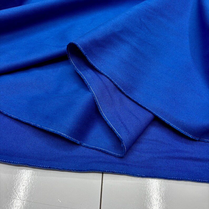 Puede incluir: Primer plano de una tela azul vibrante, posiblemente un textil o material de ropa. La tela est&aacute; doblada, revelando capas y un reverso azul m&aacute;s claro. Se ven costuras blancas en los bordes, lo que sugiere una costura terminada. El material parece liso y posiblemente tejido.