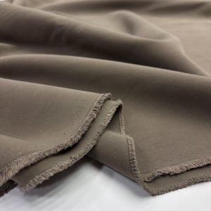 May include: A length of solid taupe-colored fabric featuring a distinct diagonal twill weave pattern. The material is draped to show soft folds and ripples, with visible frayed edges along the cut sections of the textile.