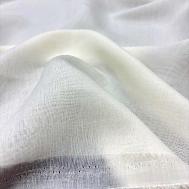 May include: A close-up shot of white, translucent fabric with a fine woven pattern. The material is arranged in gentle folds, casting shadows. The edge reveals a row of small, evenly spaced perforations, indicating a finished hem. The fabric appears delicate and light.