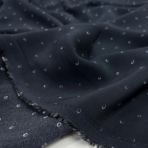 May include: Dark navy fabric featuring a repeating pattern of small, white, crescent-shaped designs. The fabric is draped, showing folds and texture. The edges are frayed, revealing the fabric's weave.
