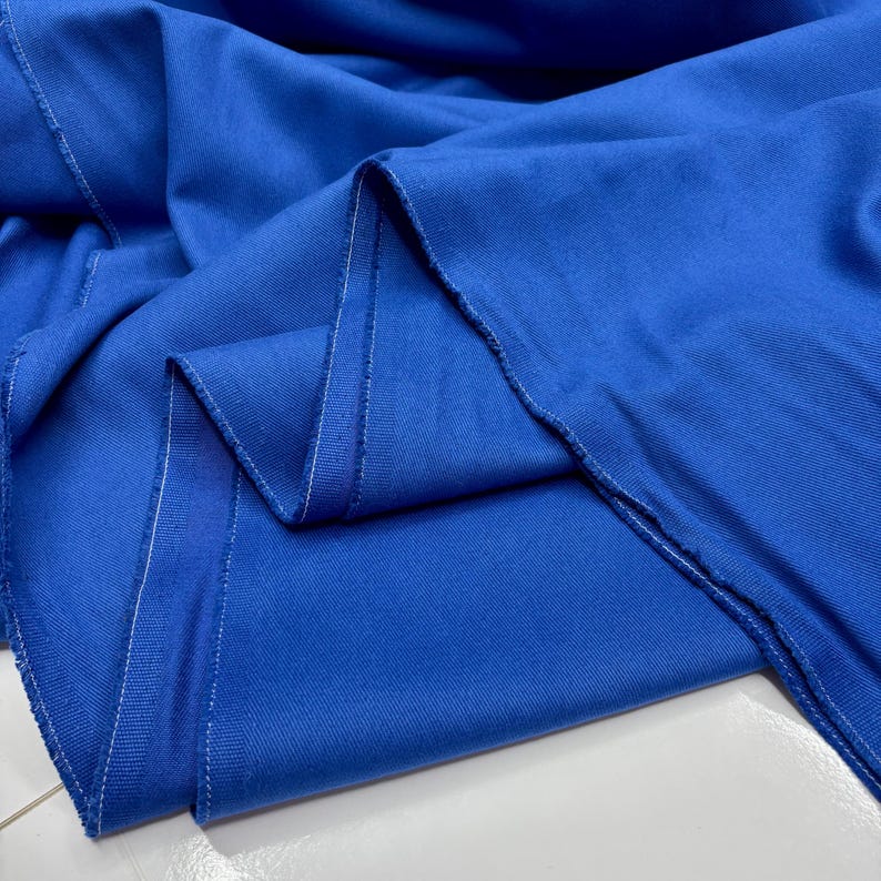 Puede incluir: Primer plano de tela azul vibrante con textura visible y costuras blancas en los bordes. La tela est&aacute; doblada, creando sombras y resaltando la ca&iacute;da del material. El color brillante sugiere su uso para ropa o decoraci&oacute;n del hogar.