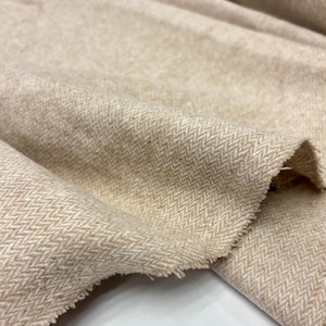 Puede incluir: Primer plano de tela de espiga beige. La tela tiene una superficie texturizada y un patrón de tejido diagonal clásico. Los bordes de la tela están deshilachados, lo que sugiere un estado en bruto o inacabado. La tela parece suave y flexible.