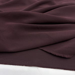 May include: A deep dark burgundy fabric with a smooth, matte texture and a fine weave. The material is draped in soft, flowing folds, showcasing the consistent solid color and the raw, slightly frayed edge along the bottom of the textile piece.
