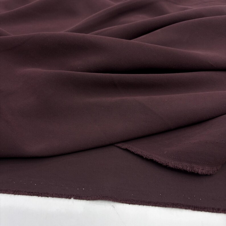 May include: A deep dark burgundy fabric with a smooth, matte texture and a fine weave. The material is draped in soft, flowing folds, showcasing the consistent solid color and the raw, slightly frayed edge along the bottom of the textile piece.