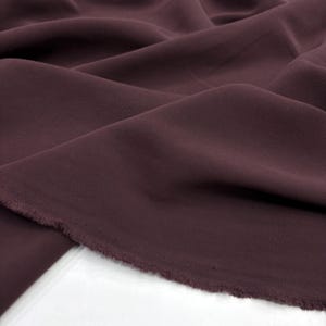 May include: A length of solid dark plum colored fabric with a smooth, matte texture. The material is draped in soft, flowing folds, showcasing its flexible quality. The bottom edge of the textile features a raw, unfinished frayed selvage border.