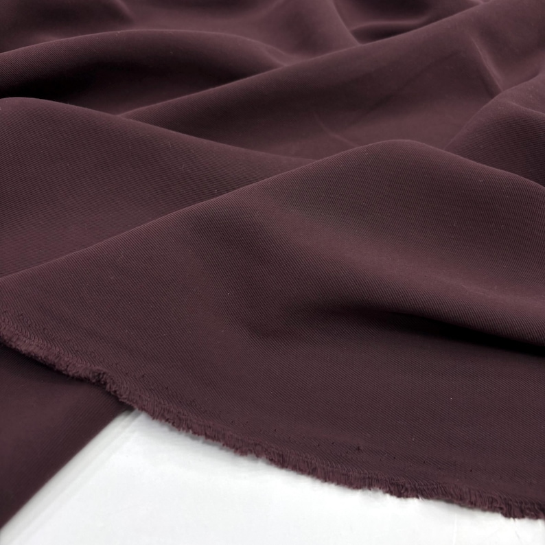 May include: A length of solid dark plum colored fabric with a smooth, matte texture. The material is draped in soft, flowing folds, showcasing its flexible quality. The bottom edge of the textile features a raw, unfinished frayed selvage border.