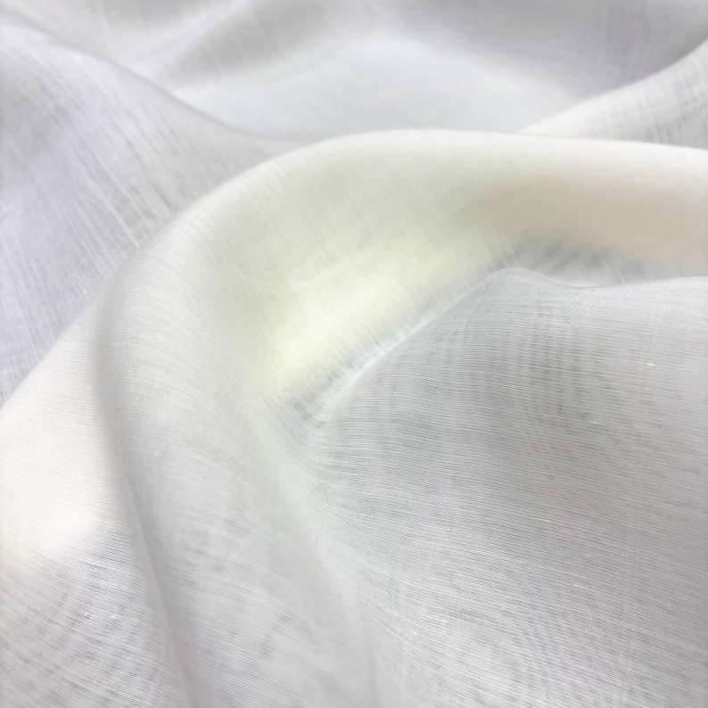 May include: Close-up of a white, sheer fabric with a subtle texture. The fabric is draped, creating soft folds and shadows. The material appears lightweight and slightly translucent, with a gentle sheen.