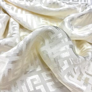 May include: Cream-coloured fabric with a repeating geometric design in a lighter, silvery hue. The fabric is draped, creating soft folds and shadows. The pattern consists of interconnected, stylised shapes.