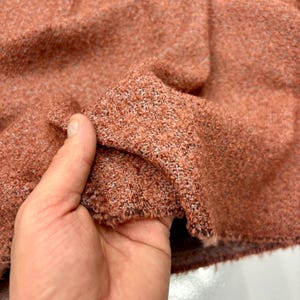 May include: Close-up of a textured fabric in a warm, reddish-brown hue. The fabric appears to be a woven material with a slightly rough texture. The edges of the fabric are visible, showing the raw, unfinished edges. The fabric is being held up.