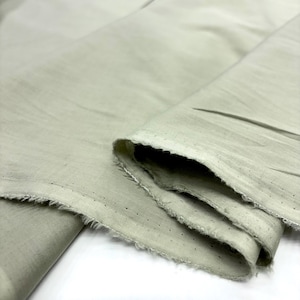 Puede incluir: Primer plano de una tela verde pálido doblada con un borde crudo y deshilachado. La tela parece ligera y tiene pequeñas perforaciones a lo largo del borde. Los pliegues crean sombras y textura, resaltando la caída del material.