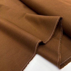 May include: Close-up of a folded piece of brown fabric. The fabric has a textured appearance with visible diagonal lines. The edges are frayed, with a white stitch along the border. The fabric is a solid, warm brown color.