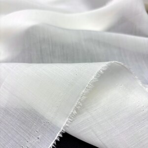 May include: Close-up of white fabric with a textured weave. The fabric is folded, creating soft shadows and highlights. The edge of the fabric is unfinished, showing a raw, fringed edge. The material appears lightweight and airy.