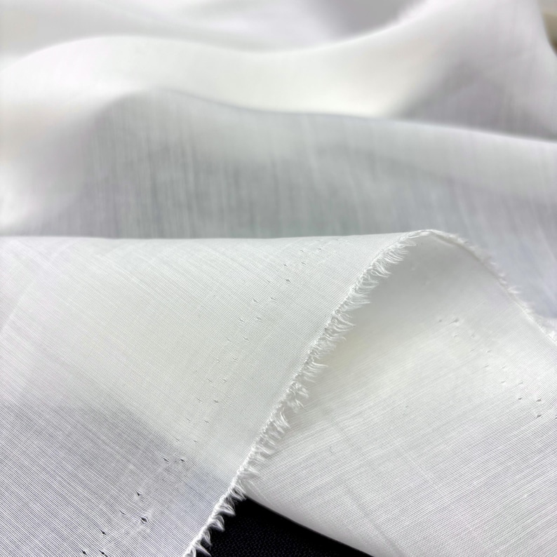 May include: Close-up of white fabric with a textured weave. The fabric is folded, creating soft shadows and highlights. The edge of the fabric is unfinished, showing a raw, fringed edge. The material appears lightweight and airy.