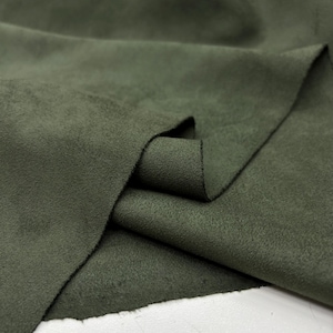 May include: Close-up of a piece of olive green suede fabric. The fabric is soft and textured, with visible folds and creases. The color is a muted, earthy tone, and the material appears to be high-quality.