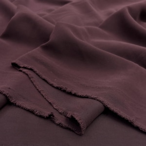 May include: A length of deep plum-colored fabric with a soft, matte texture. The material is draped in loose, flowing folds, revealing a raw, unfinished edge with visible frayed threads along the cut side of the textile piece.