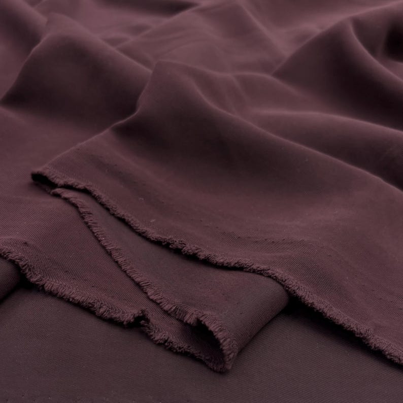 May include: A length of deep plum-colored fabric with a soft, matte texture. The material is draped in loose, flowing folds, revealing a raw, unfinished edge with visible frayed threads along the cut side of the textile piece.