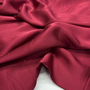 Pode incluir: Close-up de um tecido bordô profundo com uma textura suave e fluida. O material é drapeado, criando dobras e sombras suaves. A cor é um tom vermelho rico e saturado, com um leve brilho.