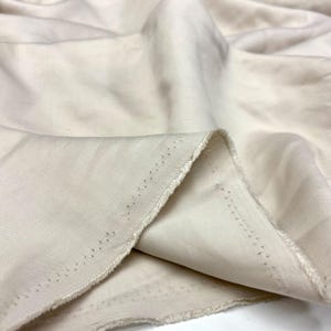May include: A length of off-white fabric with a visible diagonal twill weave pattern. The material features soft, flowing folds and drapes, with a raw, unfinished edge showing a line of small, evenly spaced pinholes along the selvage border.