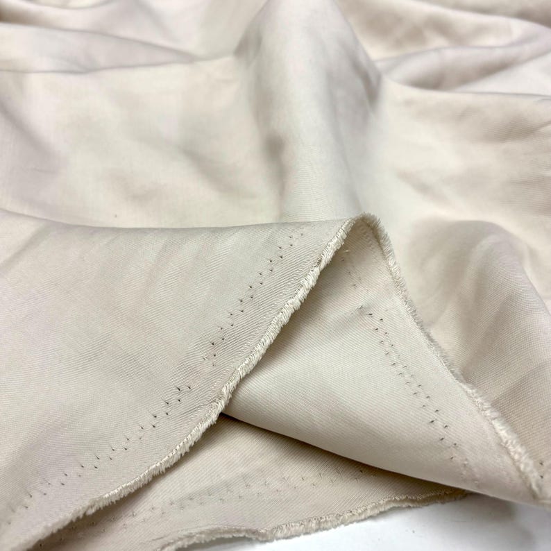 May include: A length of off-white fabric with a visible diagonal twill weave pattern. The material features soft, flowing folds and drapes, with a raw, unfinished edge showing a line of small, evenly spaced pinholes along the selvage border.