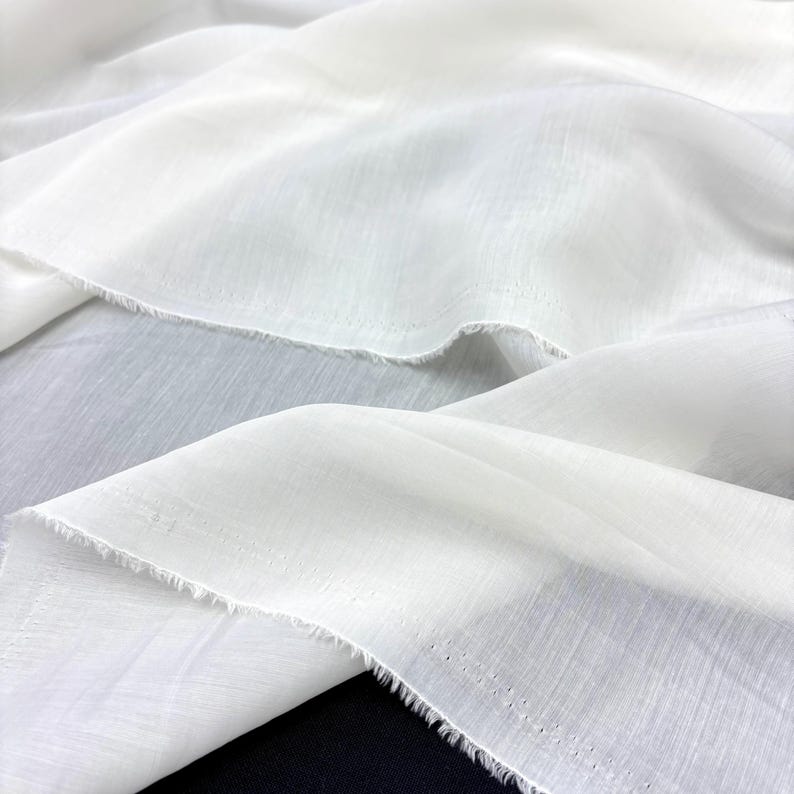 May include: Close-up of white fabric with a delicate, semi-transparent texture. The fabric is draped in soft folds, creating shadows and highlights. The edges of the fabric have a raw, unfinished look. The overall effect is one of lightness and elegance.
