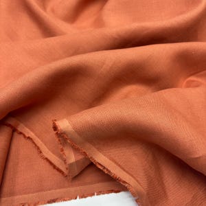 May include: Close-up of a folded piece of terracotta-colored linen fabric. The fabric has a textured appearance with visible weave details. The edges of the fabric are frayed, indicating a cut edge. The overall tone is warm and earthy.