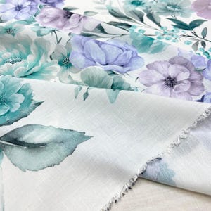 May include: Close-up of white fabric with a floral pattern. The design features large, watercolor-style flowers in shades of teal, lavender, and purple, with green leaves. The fabric has a slightly textured appearance and a raw edge.