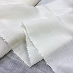 May include: Close-up of white fabric with a slightly textured weave. The edges are frayed, and the fabric is folded, creating shadows and depth. The material appears lightweight, possibly linen or cotton.